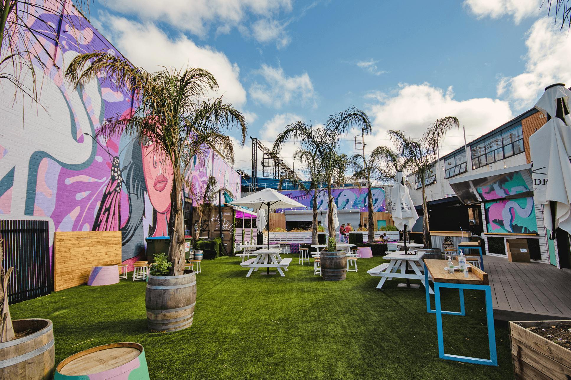 An outdoor garden bar with palm trees, lawn, and pink coloured art murals.